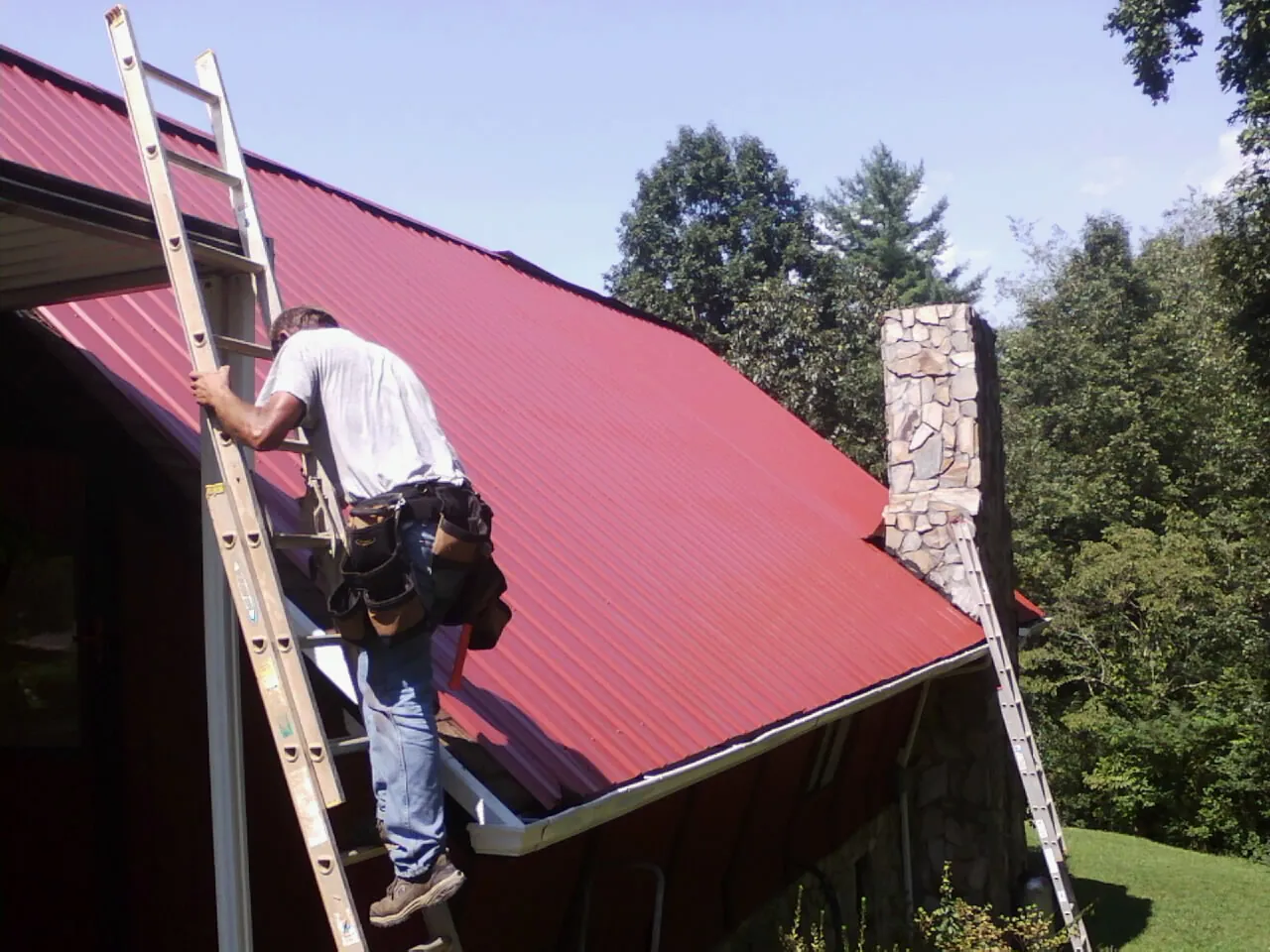 Roofer on a ladder beside a red metal roof and stone chimney — Soffit & Fascia Repair by Roofing Brothers in Bunch